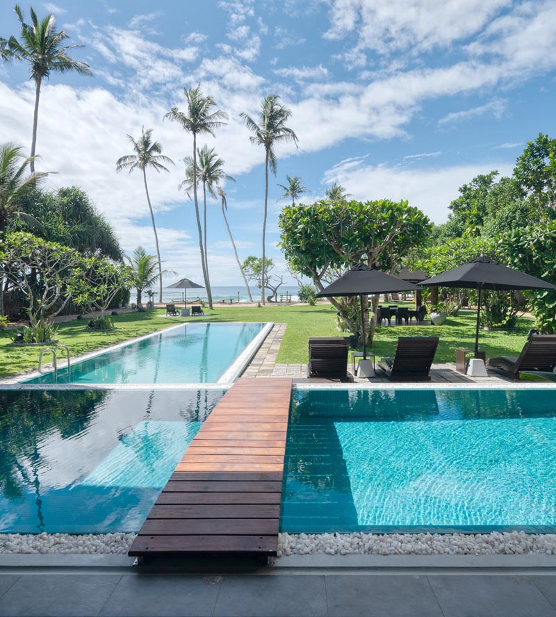 The two pools at Mirissa Beach Villa joined by a wooden walkway, with the ocean and palms beyond