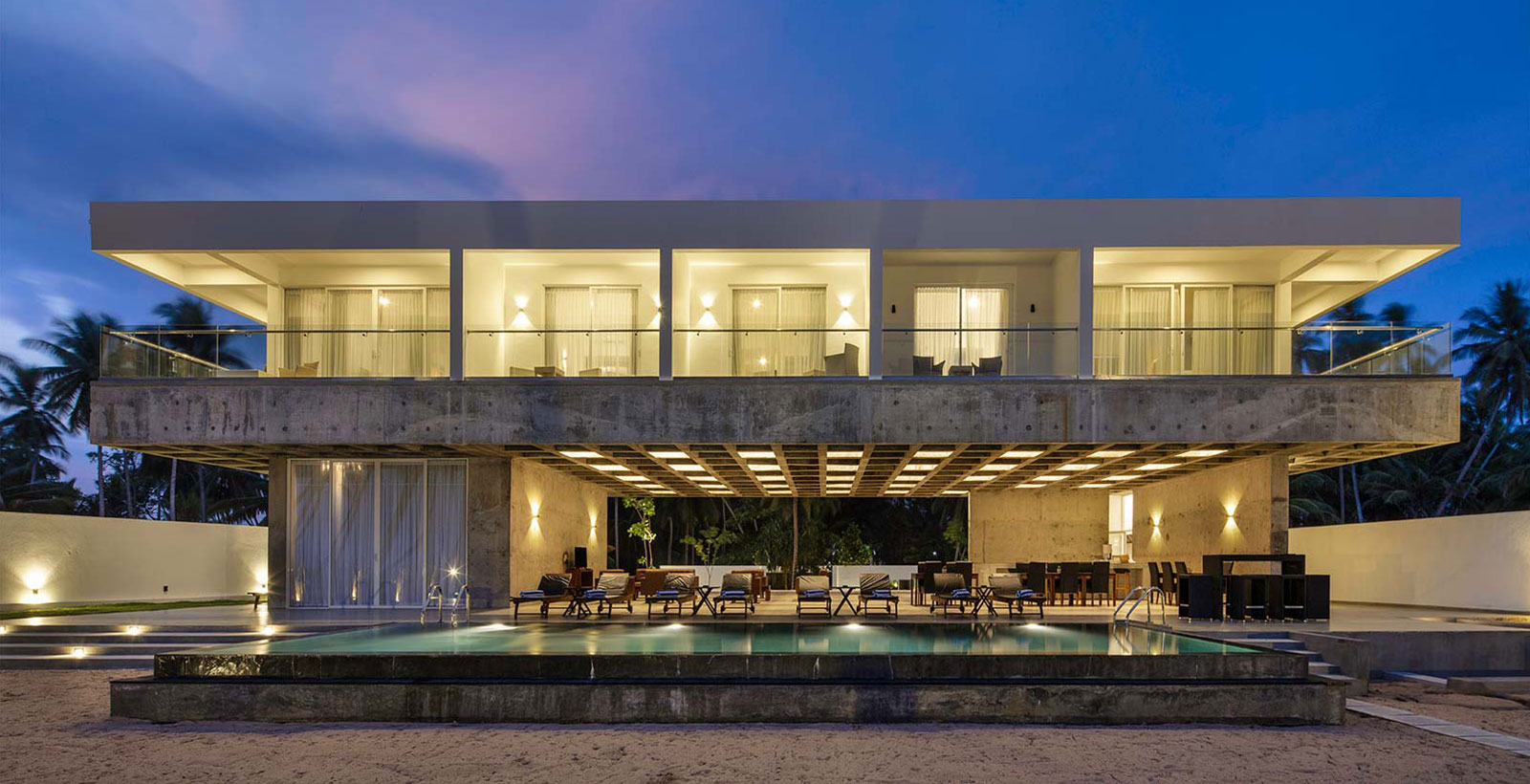 Beachfront villa and long pool at dusk, rooms lit, loungers on the deck